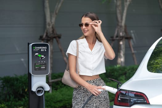 Young woman travel with EV electric car charging in green sustainable city outdoor garden in summer. Urban sustainability lifestyle by green clean rechargeable energy of electric BEV vehicle innards