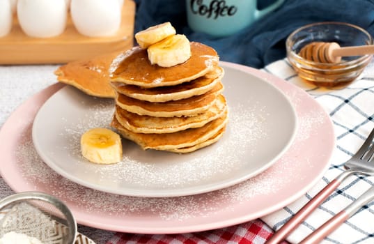 homemade banana pancakes on plate sprinkled powder and honey. Pancakes on the kitchen table against the background of eggs and cups with coffee. Pancake slide with honey topping and fresh bananas