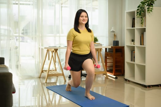 Overweight young woman doing lunges exercise with dumbbells on mat in living room.