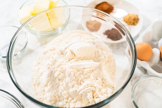 Overhead view of a glass mixing bowl filled with flour, with key gingerbread spices, butter, and eggs arranged in the background. These ingredients will be combined to create the Classic Gingerbread Cookie Dough, perfect for holiday baking.
