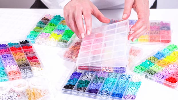 Womans hands gracefully poised over a collection of beads, sorted by color in transparent organizers. The array of beads spans a vibrant spectrum, from deep purples to bright oranges, meticulously arranged for easy selection as she embarks on creating a custom piece of jewelry.
