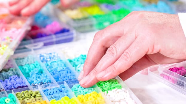 Womans hands gracefully poised over a collection of beads, sorted by color in transparent organizers. The array of beads spans a vibrant spectrum, from deep purples to bright oranges, meticulously arranged for easy selection as she embarks on creating a custom piece of jewelry.