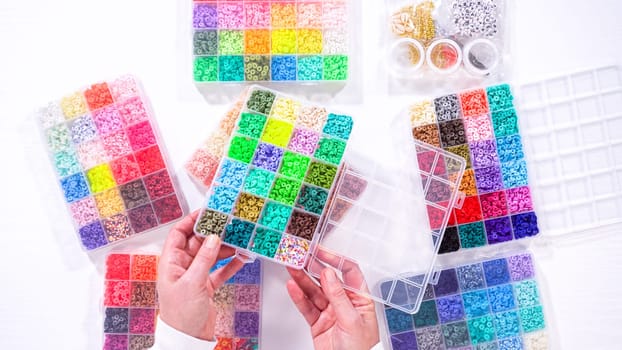 Flat lay. Womans hands gracefully poised over a collection of beads, sorted by color in transparent organizers. The array of beads spans a vibrant spectrum, from deep purples to bright oranges, meticulously arranged for easy selection as she embarks on creating a custom piece of jewelry.
