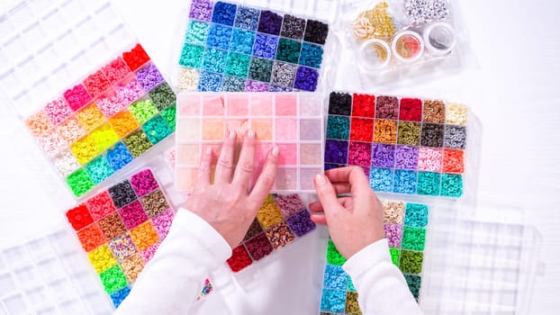 Flat lay. Woman hands gracefully poised over a collection of beads, sorted by color in transparent organizers. The array of beads spans a vibrant spectrum, from deep purples to bright oranges, meticulously arranged for easy selection as she embarks on creating a custom piece of jewelry.