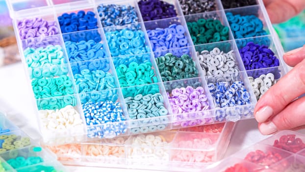 Woman hands gracefully poised over a collection of beads, sorted by color in transparent organizers. The array of beads spans a vibrant spectrum, from deep purples to bright oranges, meticulously arranged for easy selection as she embarks on creating a custom piece of jewelry.