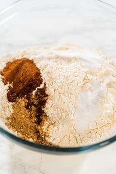 Close-up view of a large glass mixing bowl with flour and a measured amount of spices (including cinnamon and ginger) added on top, ready to be mixed for the Amber Gingerbread Cookie Dough.