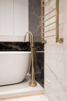 Long stylish Gold faucet on the floor near white bathtub on granite wall background. Modern bathroom interior.