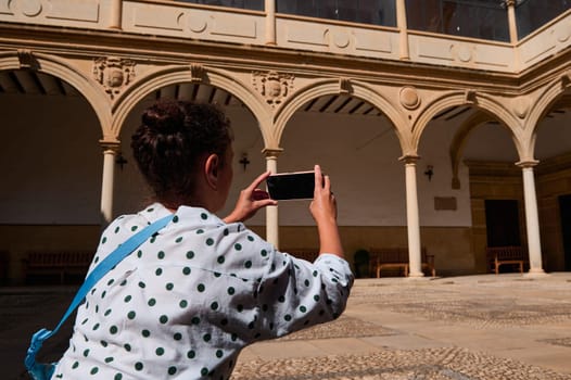 A female tourist captures the beauty of historical architecture in a sunny courtyard using her smartphone, highlighting travel and exploration.