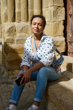 A woman in polka dot shirt and jeans sits on ancient stone steps, holding a phone, basking in the sunlight. Her relaxed expression suggests a moment of leisure and reflection.