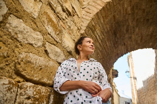 A woman in a polka dot shirt stands against a stone wall under an archway, enjoying an outdoor exploration in a historic location.