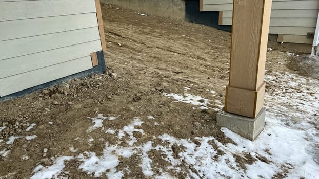 Close-up view of a house foundation surrounded by dirt and light patches of melting snow. Wooden posts and siding are visible, suggesting construction or a backyard area in winter conditions.
