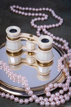 Binoculars rest on mirror surrounded by pearl necklaces. The image evokes elegance and timelessness.