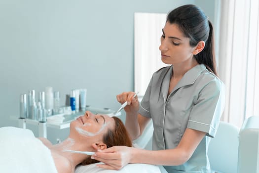 Professional masseuse applying face mask with brush on woman's face at cosmetology salon. Relaxed female customer is getting pampered in spa by beautician.