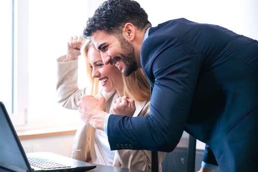 Male and female colleagues in formal attire cheerfully celebrating a victory while looking at a laptop. Bright and modern office setting reflects their enthusiasm and teamwork.