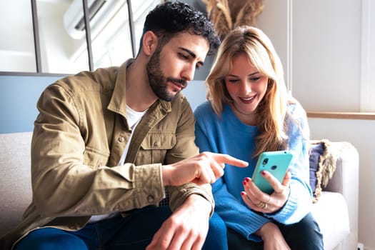 Young man and woman sitting on a couch, sharing a moment of excitement while looking at a smartphone. Casual outfits and a relaxed indoor environment.