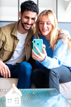 A joyful couple sits on a cozy couch, smiling and waving at a smartphone during a video chat. Their happy expressions and relaxed setting highlight connection, technology, and relationships.Vertical