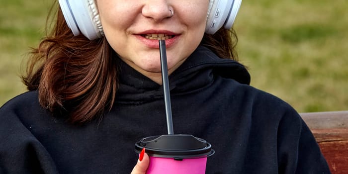 A close-up shot of a young woman wearing headphones, sipping from a pink cup with a black straw. She is sitting outdoors on a bench, with green grass in the background.