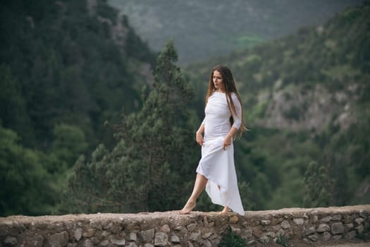 A woman in a white dress is standing on a stone wall in the mountains. She is alone and is looking out into the distance. The scene has a serene and peaceful mood