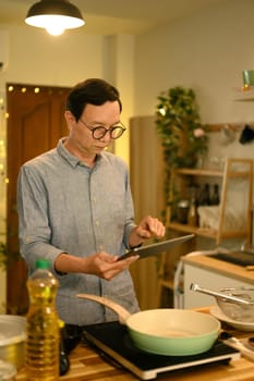 Portrait of senior man standing in warm kitchen, reading online recipe on digital tablet.