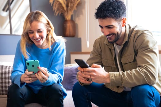 A joyful young couple sitting indoors, both holding smartphones and laughing together. They share a lighthearted moment in a modern, cozy living room setting.