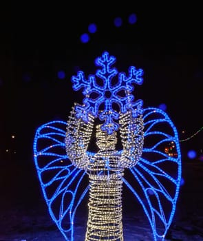 An enchanting angel figure adorned with blue lights stands gracefully at night, holding a large snowflake, enhancing the winter ambiance.