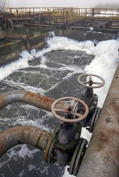 Valves are seen controlling the flow of water with foam forming in the treatment basin at an old facility during daytime.