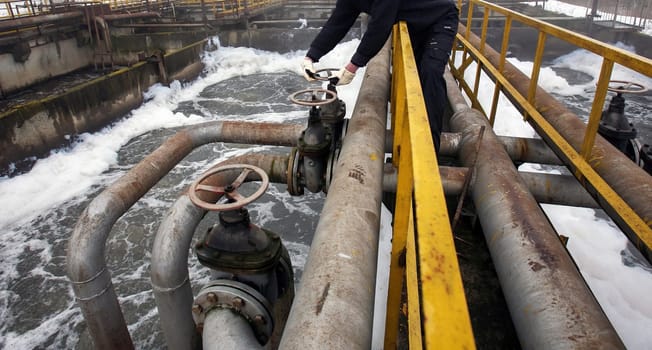 A worker adjusts valves on large pipes at an old water treatment facility while water flows through the system, highlighting maintenance efforts.
