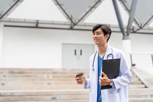 A male doctor in a white coat stands outside a hospital, holding a clipboard and coffee, exuding professionalism and confidence.