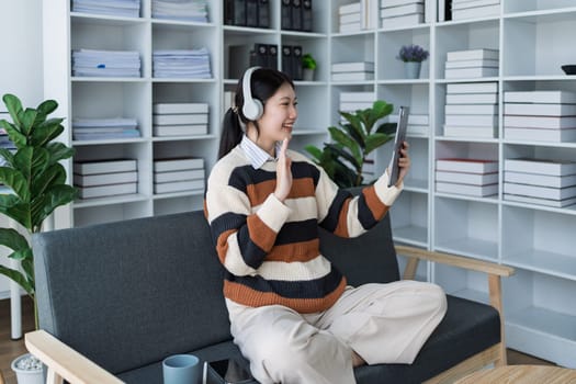 A cheerful woman enjoys a video call using a tablet while sitting comfortably on a couch in a modern office setting, surrounded by plants and organized shelves.