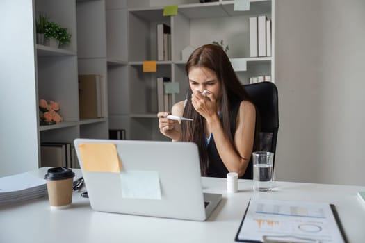 Young woman in office sneezing, showing symptoms of a cold, surrounded by work materials.