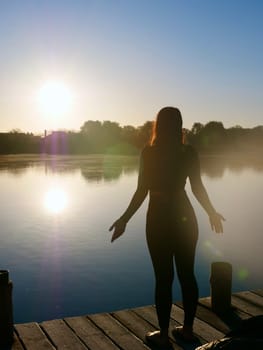 Woman standing on sadhu board, process of standing barefoot on nails, yoga practice and meditation outdoors, autumn day near lake at sunrise