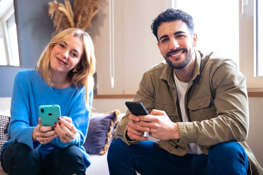 A cheerful young couple sitting indoors, both holding smartphones and smiling warmly. The woman leans slightly, creating a relaxed and friendly atmosphere in a cozy setting.