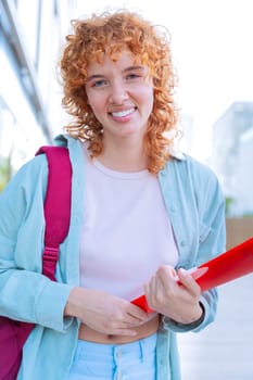 A cheerful young red-haired woman wearing a casual outfit and a backpack holds books and a smartphone. She stands outdoors in a bright urban environment, exuding confidence and positivity.