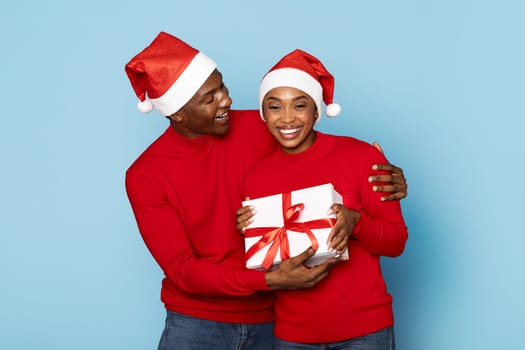 A joyful couple wearing red sweaters and Santa hats smiles while exchanging a beautifully wrapped gift. The festive atmosphere highlights the spirit of giving during the holidays.