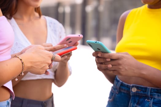Three cheerful young women gathers around a red smartphone, reacting with excitement. They are dressed casually, standing outdoors in a modern urban environment, enjoying a fun moment together.