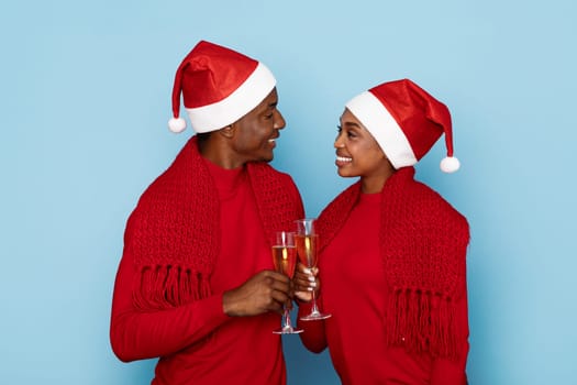 A joyful couple in red outfits, adorned with Santa hats, clinks champagne glasses together in a festive celebration. Their bright smiles radiate holiday cheer against a light blue background.