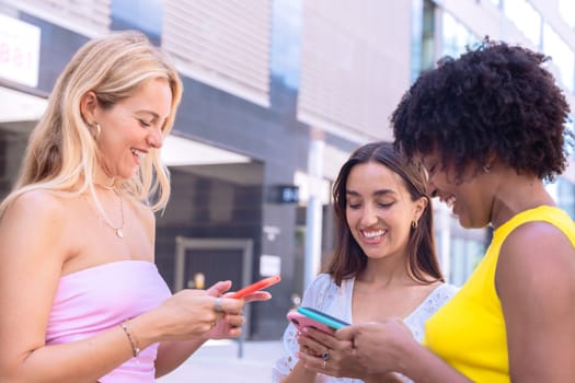 Three cheerful young women gathers around a red smartphone, reacting with excitement. They are dressed casually, standing outdoors in a modern urban environment, enjoying a fun moment together.