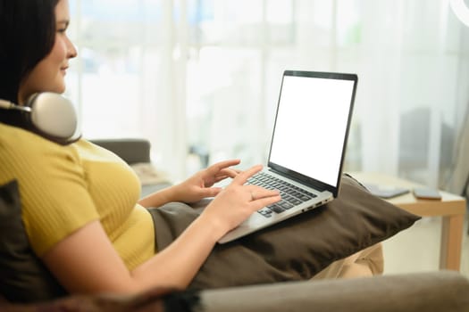 Side view of smiling young woman in casual clothes using laptop in bright living room.
