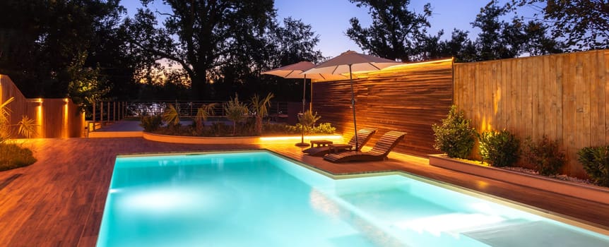A nighttime scene of a pool and deck illuminated with warm lighting, surrounded by lush greenery, creating a luxurious and tranquil setting. Concept of relaxing outdoor atmosphere and elegant evening leisure