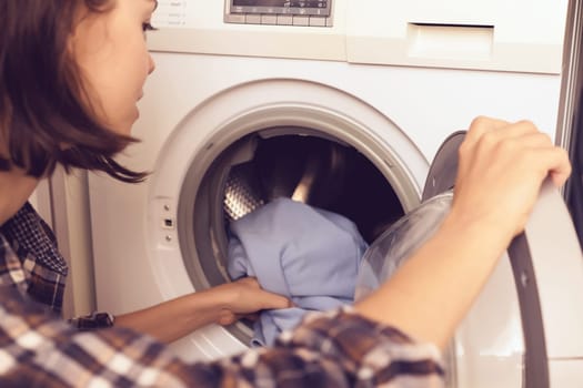A girl folds bright clothes into a new washing machine in the bathroom at home, a woman washes things and spin them out with detergent in the laundry close-up, top view.