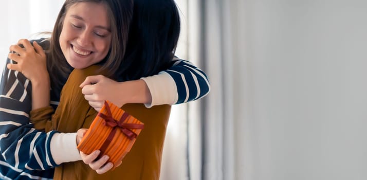 Two happy young girls laugh, friends hug and give each other gifts during the holiday. Women surprise and congratulate on the anniversary of their relationship in their cozy home.