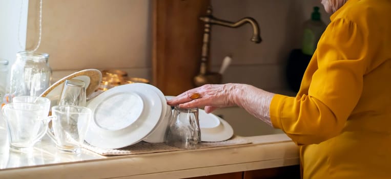 Elderly retired gray-haired woman having a good time in her house, active in her old 80+ age, washing dishes in a cozy kitchen, cleaning. A happy grandmother in a yellow blouse, young at heart.