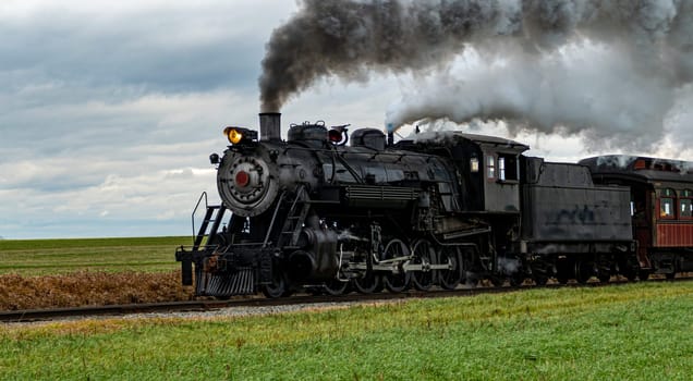 A vintage steam train chugs along railway tracks in a vast rural area, emitting thick plumes of smoke against a backdrop of dark clouds. Nearby fields exhibit golden crops.