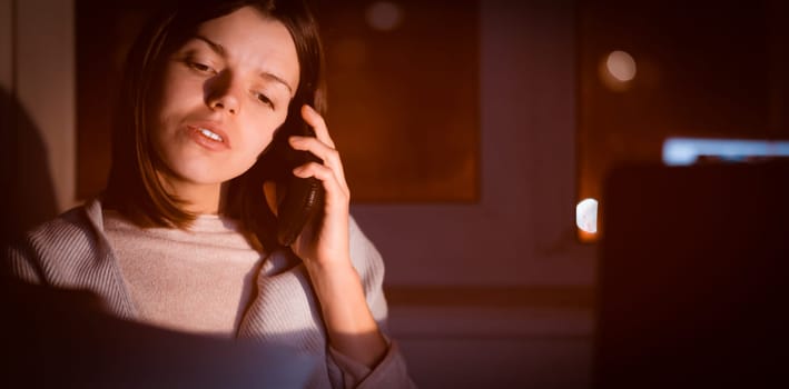 A young girl works at home with a laptop at night and talks on the mobile phone, goes through papers, documents, compiles analytics, makes calculations and notes, solves business issues, studies.