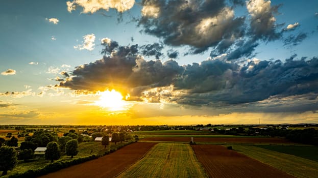 A vibrant sunset casts warm colors over expansive farmland, with lush green fields and patches of clouds creating a picturesque sky. The serene landscape reflects the tranquility of rural life.