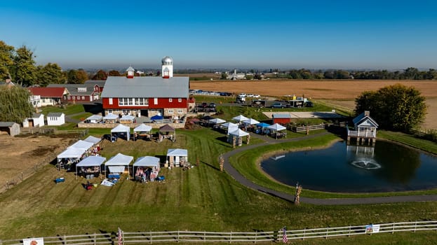 Ronks, Pennsylvania, USA, October 20 2024 - Visitors enjoy a lively autumn market set in front of a large red barn, featuring white tents and a serene pond. The clear blue sky enhances the cheerful ambiance of the day.