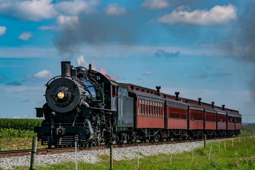 A classic steam train chugs along the tracks, billowing smoke into the sky. Surrounded by green fields, the vintage train transports passengers, evoking nostalgia.