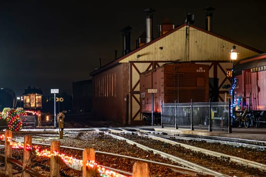 Strasburg, Pennsylvania, USA December 7, 2024 - A classic steam locomotive steams beside a festive station, adorned with colorful lights for the holiday season.