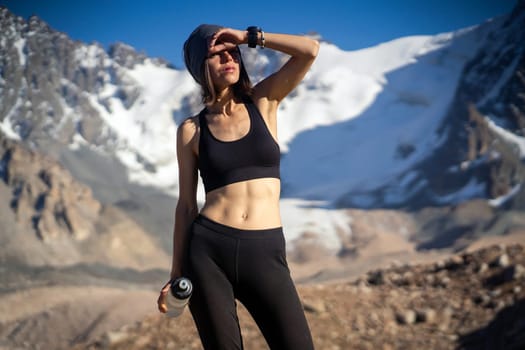 A young athletic girl drinks water, isotonic from a bottle during training, trail running in the snow-capped mountains. Runner is making workout outdoor.