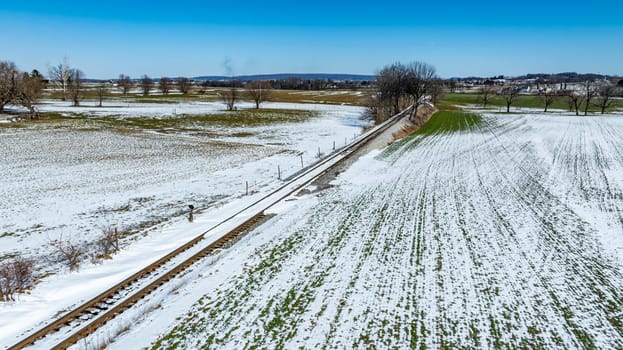 A vast expanse of snow-covered fields stretches across the rural landscape, with a railway track running alongside. The clear blue sky above enhances the winter scenery, revealing distant hills.